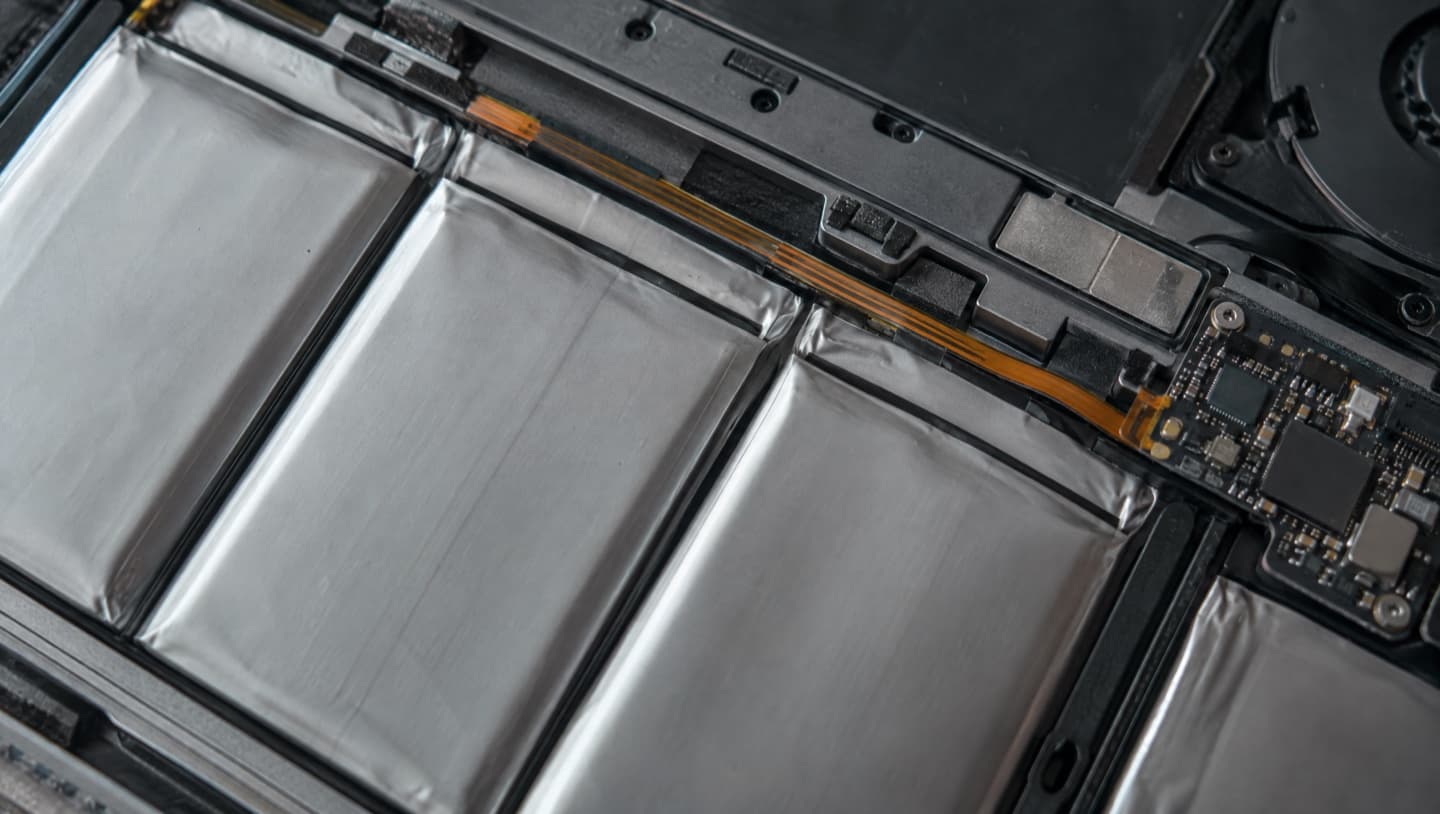 Close-up of the flat silver pouch cells of a laptop battery inside an opened laptop, with its connector ribbon and the small control board at one end.
