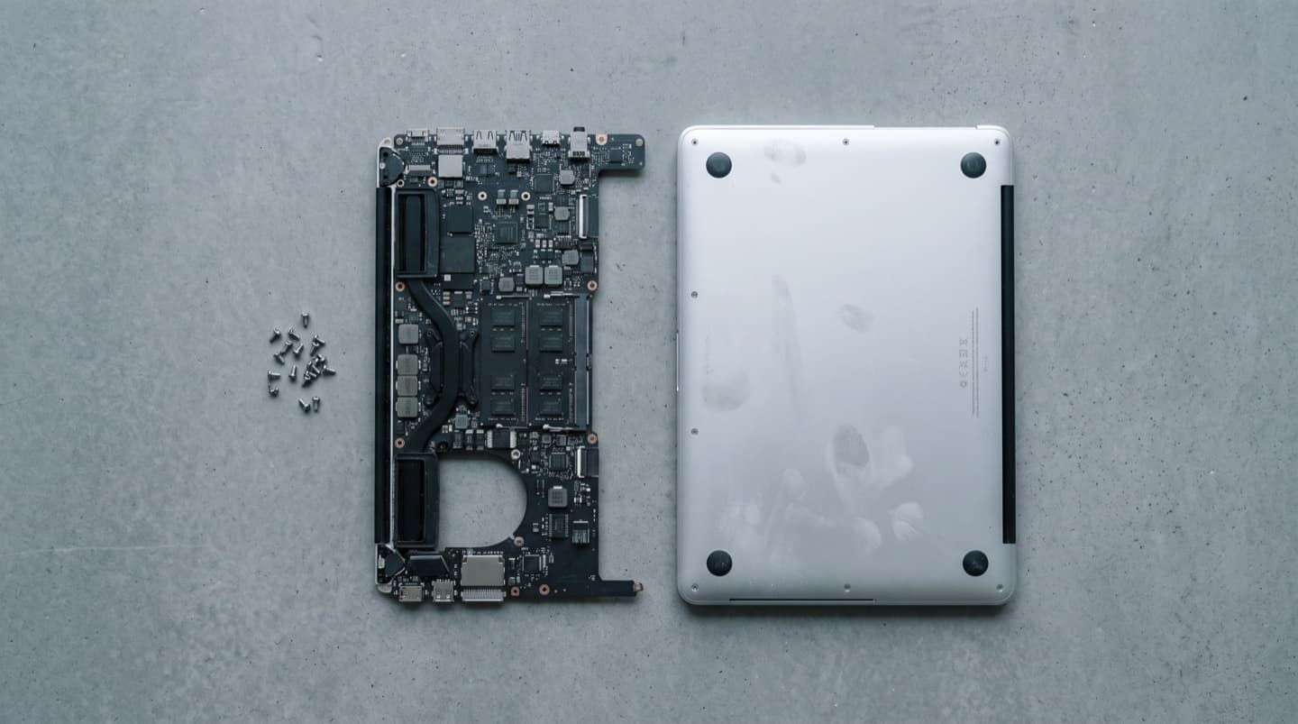 A laptop logic board lifted clear of its case, laid beside the machine's aluminium bottom panel and its screws, shot from directly above.
