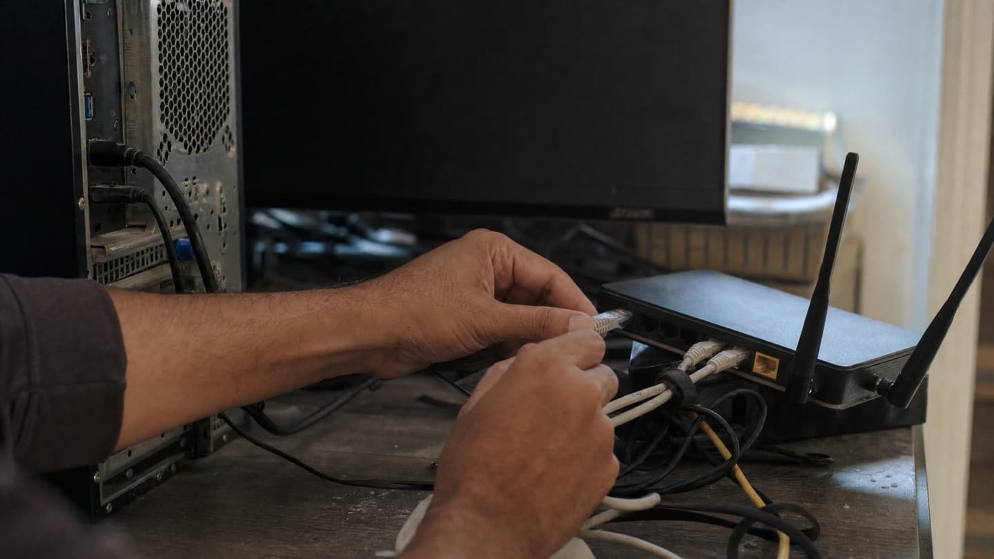 A technician's hands checking a router and cables next to a desktop tower in a home office.