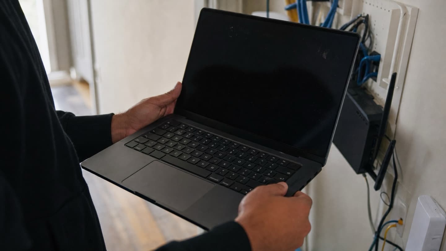 A laptop held near a hallway wall to check Wi-Fi signal, cables and a router visible in the background.