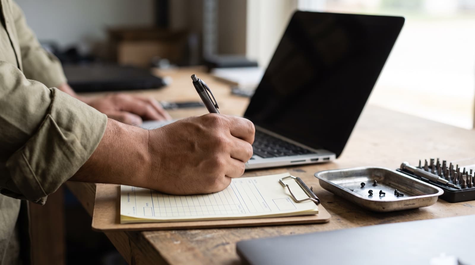 Technician filling in a paper work ticket beside an open laptop
